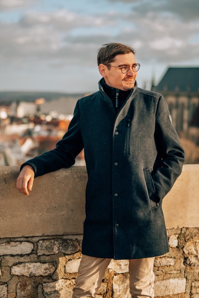 Mann mit Sonnenbrille und dunklem Mantel vor einem Stadtpanorama